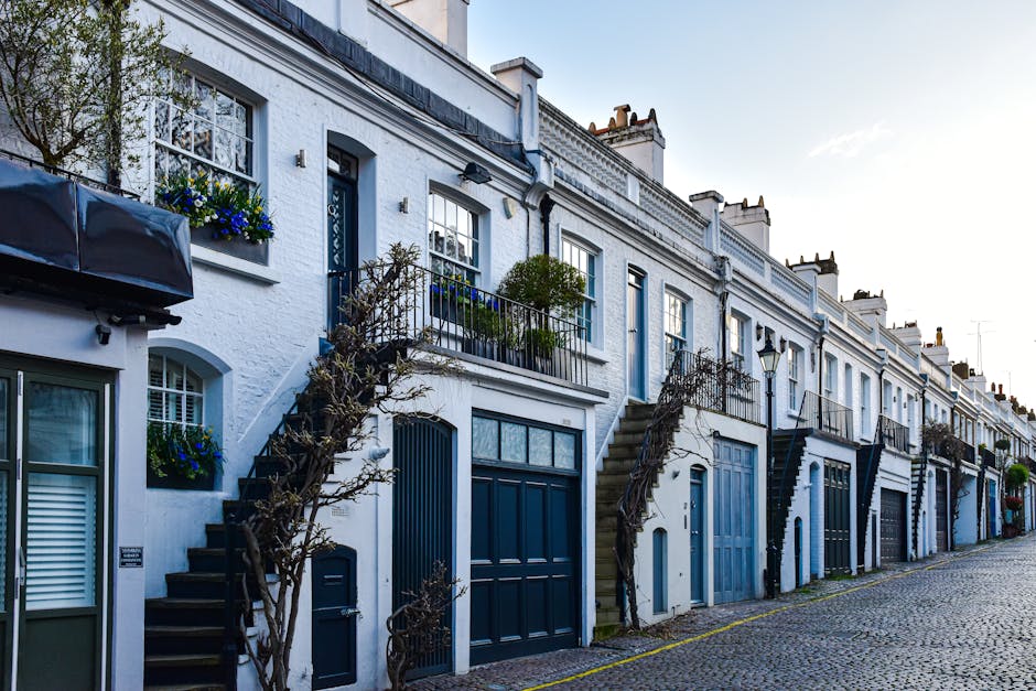 A row of white terraced houses with modern blue garage doors and exterior staircases leading to upper-floor entrances, situated on a cobbled street during daylight. Each property features decorative w