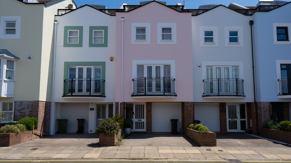 A row of white terraced houses with modern blue garage doors and exterior staircases leading to upper-floor entrances, situated on a cobbled street during daylight. Each property features decorative w