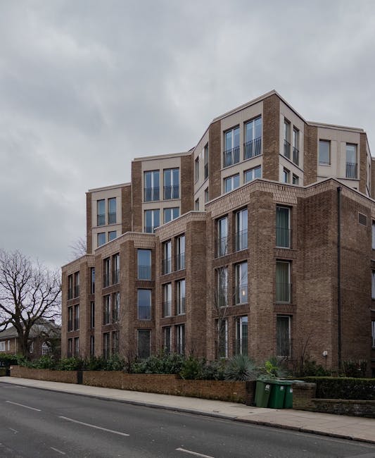 A large multi-storey residential building with a modern architectural design, featuring a combination of brown brick and light-colored paneling, large rectangular windows, and some with metal railings
